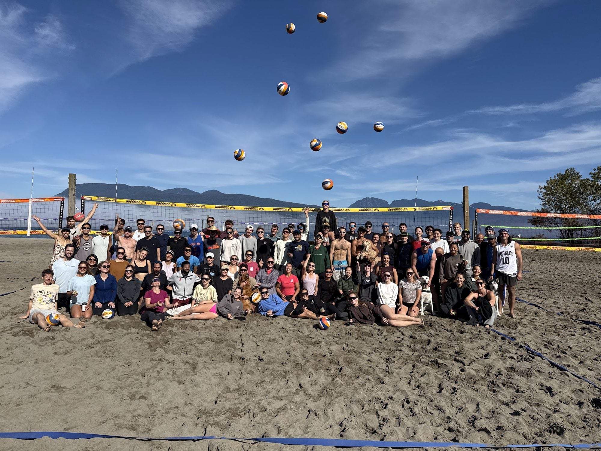 Over a hundred Never Not Sandy players posing at Spanish Banks with volleyballs thrown in the air